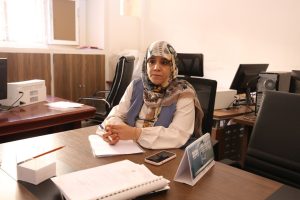 2B83311D-CE71-43BA-B292-243117011A2A - جامعة سبها | Sebha University Woman wearing a patterned hijab sits at an office desk with a notebook, pen, and smartphone, facing the camera.