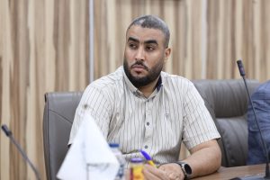 2BC167AA-74FF-4C8B-9B07-BD5BE60D7768 - جامعة سبها | Sebha University Man with a beard in a striped button-up shirt sits at a conference table, looking to the side in a meeting room.