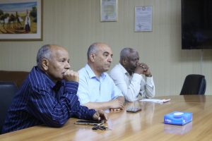 2D408056-B589-49E6-A67C-44C39C9D055D - جامعة سبها | Sebha University Three men sit at a wooden conference table, hands on chins, appearing thoughtful during a meeting in an office setting with phones and a tissue box on the table.