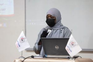 2E6F2895-9C50-4A74-983A-6183F29F88AD - جامعة سبها | Sebha University Woman in a gray hijab and black mask speaks into a microphone at a desk with a laptop and small flags nearby.
