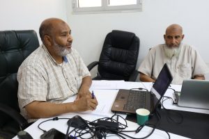 Two men sit at a white conference table with laptops, papers, and a paper cup, engaged in a discussion in an office.