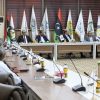 Formal diplomatic meeting in a flag-lined conference room, delegates seated around a long table with microphones and documents in front of them.