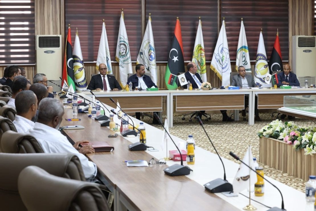 Formal diplomatic meeting in a flag-lined conference room, delegates seated around a long table with microphones and documents in front of them.