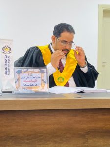 35588D1D-3838-42BA-BF05-A4352DDA876F - جامعة سبها | Sebha University Graduate in black gown and yellow stole sits at a desk, adjusting glasses while signing documents; signs and university banner visible nearby.