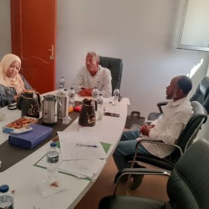 397574FE-02E1-4E02-906D-03E6214C7EE9 - جامعة سبها | Sebha University Three people sit around a conference table in a meeting room, with papers, water bottles, and snacks on the table as they discuss.','A man in a light shirt speaks while two others listen, at a white-clothed meeting table with beverages and documents nearby.','Woman wearing a hijab and two men are seated around a rectangular table with folders, water bottles, and pastries visible.','Office meeting scene: documents and notebooks spread across the table, several water bottles and coffee flasks, and three people engaged in discussion.','Group of colleagues in a conference room, discussing a document-filled table with drinks and snacks nearby.