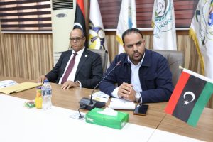 407F8773-1421-49BE-8883-118F089AE5A7 - جامعة سبها | Sebha University Two men sit at a conference table in a formal meeting; one in a suit, the other in a blue jacket, with microphones and flags in the background and Libyan flag on the table.