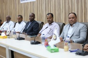 Five men sit at a long conference table with microphones, flags, and bottled drinks during a formal meeting in a boardroom.