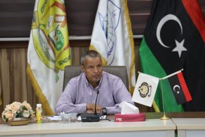 A man seated at a conference table with microphones, flags, and flowers in the background.