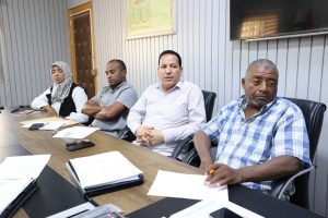 4473AEF9-1A2A-4FA3-9317-1D269360BA18 - جامعة سبها | Sebha University Four people sit around a conference table with papers and notebooks in a meeting room, listening attentively.