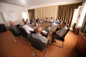 46455D09-2B42-493A-8E0B-0E0E0C33F114 - جامعة سبها | Sebha University A group of professionals seated around a large rectangular conference table in a meeting room, with notebooks, bottles of water, and a whiteboard on the wall.