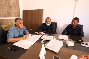 46DFEDE1-AE9D-437C-860F-67B49A798C47 - جامعة سبها | Sebha University Three men sit at a cluttered office table reviewing documents in a meeting room.