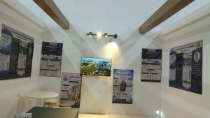 48C4B2B7-951C-4383-B3DB-0E7F88B6F22B - جامعة سبها | Sebha University Exhibition room with framed posters on cream walls, a wall-mounted TV showing a landscape, and ceiling track lighting.