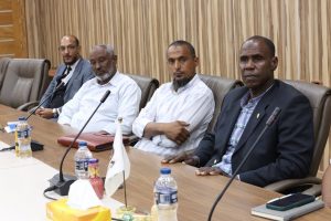 48D37BAE-211D-47B9-A86E-D0FD223BAF56 - جامعة سبها | Sebha University Four men in business attire sit along a conference table with microphones, water bottles, and a tissue box in a boardroom setting.