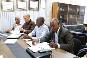 48ECBDBB-C592-42BA-BBC8-2BF505A8DB3D - جامعة سبها | Sebha University Four men sit at a conference table reviewing documents in a formal office with framed certificates on the wall and a glass-front cabinet in the background.