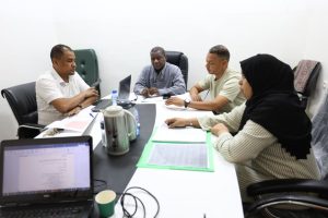 49EE138A-75F1-4F76-B465-A33D00FAD9C9 - جامعة سبها | Sebha University A group of five professionals sit around a conference table, discussing with laptops, papers, and a thermos at hand.
