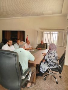 Six people seated around a conference table in a bright meeting room, discussing documents and notes on the table.