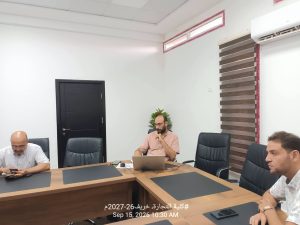 52FDF2F1-A4F8-4556-8A37-36A5665DC959 - جامعة سبها | Sebha University Conference room scene with four men seated around a large wooden table, using laptops and phones; one man in a pink shirt works on a laptop while others look at devices. White walls, a black door, striped window blinds, and a wall-mounted air conditioner. A date/time overlay at the bottom reads Sep 15, 2026 10:30 AM.