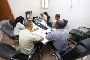 577A1E94-7E01-4E20-9A4E-D554E9ECBAC6 - جامعة سبها | Sebha University Six professionals sit around a white conference table, reviewing papers and using laptops in a meeting.