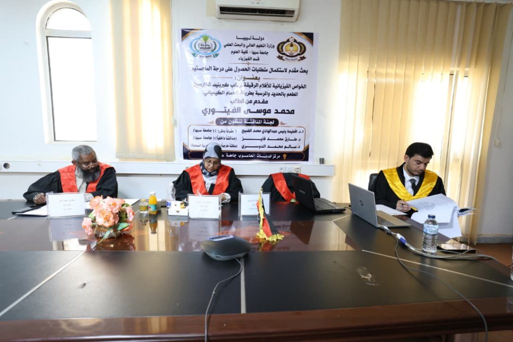 577BCFE9-564A-419A-BA86-10569A5E90F3 - جامعة سبها | Sebha University Three academics in robes sit at a conference table during a formal ceremony, with a banner and window blinds in the background.
