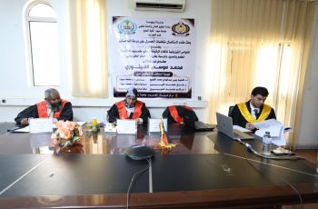 Three academics in robes sit at a conference table during a formal ceremony, with a banner and window blinds in the background.