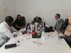 5941ED38-F849-4DBF-8391-3C254EDD32E4 - جامعة سبها | Sebha University Five professionals sit around a conference table in a meeting room, with water bottles, notes, and snacks on the table.