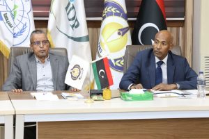 5993A5AB-8988-4D61-B6E1-C4EF893229AB - جامعة سبها | Sebha University Two men in suits seated at a conference table during a formal meeting, with flags and documents between them and a water bottle visible on the right.