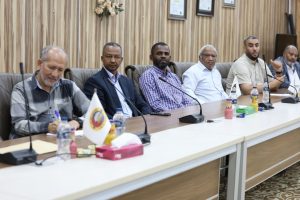5CFCAAF2-09BD-424D-AD29-1BB268DD5EC2 - جامعة سبها | Sebha University Seven men sit along a long conference table in a meeting room, with microphones, a small flag, and bottles of water and juice on the table.