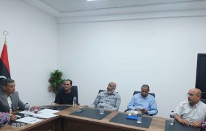 5E7E97BF-D382-49EA-B239-5F2F9E38025F - جامعة سبها | Sebha University Five men sit around a conference table in a white-walled room, discussing documents with water bottles nearby.