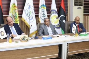 Three men in suits sit at a long conference table with flag banners in the background, papers and beverages on the table.