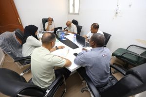5F7433F4-0312-40A9-AE4C-0908BE7DCBA2 - جامعة سبها | Sebha University A diverse group of six people seated around a white conference table, discussing documents with laptops and notebooks in a bright office.