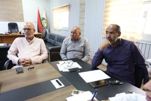 6016AAB1-FFAC-41BE-B6E1-A5ECFF2B3C15 - جامعة سبها | Sebha University Three men sit at a conference table in a meeting room, looking attentive, with papers and phones on the table and flags in the background.