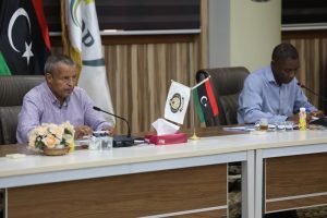 Two men sit at a conference table speaking into microphones, with Libyan and organizational flags in the background.