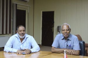 6495B600-030C-4B26-8D92-71A4E1244D83 - جامعة سبها | Sebha University Two men sit at a wooden table in a meeting room, smiling slightly and facing the camera.