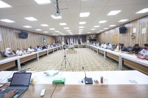 Wide view of a formal meeting with people seated around a U-shaped table in a conference room, flags at the far end and a tripod camera in the center.