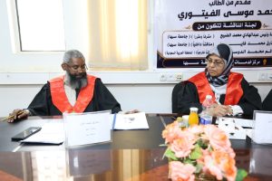 Two judges in black robes with red sashes sit at a conference table, reviewing documents as a banner in Arabic hangs behind them.