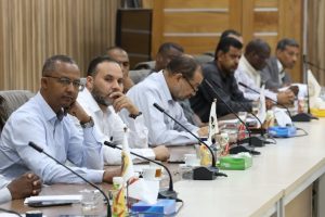 A group of men seated at a long conference table with microphones, discussing during a formal meeting.