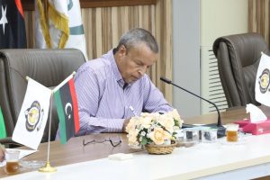 A man in a striped shirt signs a document at a conference table with flags, a floral arrangement, and a microphone nearby in a formal meeting room.
