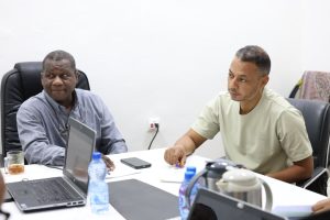 6D2B5C7C-DEDC-46B0-A020-84717B323731 - جامعة سبها | Sebha University Two men sit at a white conference table with laptops; one on the left watches while the other on the right writes with a pen.