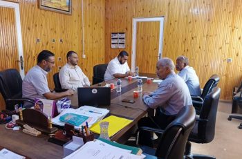 Six men sit around a large wooden conference table in a wood-paneled office, laptops, papers, and water bottles visible as they discuss and review documents.