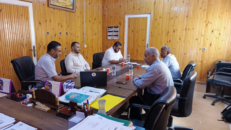 6EAD6C1D-DF7E-48F2-B034-0D7064E8CA21 - جامعة سبها | Sebha University Six men sit around a large wooden conference table in a wood-paneled office, laptops, papers, and water bottles visible as they discuss and review documents.