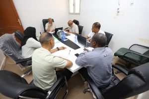 Six people sit around a rectangular conference table in a bright room, discussing with laptops, papers, and water bottles on the table.