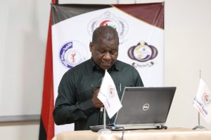 761F93B9-E38A-4D7A-AD94-A2160DA1A99E - جامعة سبها | Sebha University A man at a podium giving a presentation, looking down at notes with a Dell laptop in front of him and conference banners behind him.
