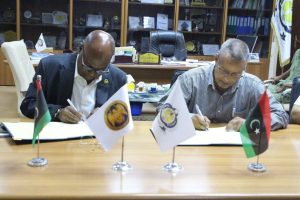 Two men sign documents at a conference table, flanked by three small flags in a formal meeting room.