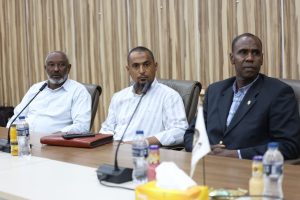 7A9C84C6-E138-49E3-8DC9-E544E7234761 - جامعة سبها | Sebha University Three men sit at a conference table with microphones, posing for a formal meeting; water bottles and a tissue box are on the table.