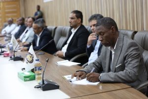 7CCC937A-4EBF-41EF-BEBD-066E2852B31C - جامعة سبها | Sebha University Man in a gray suit signs a document at a long conference table, with colleagues seated behind him in a boardroom setting.