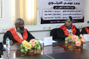 Two panelists in black robes with red sashes sit at a conference table, with a large Arabic banner behind them announcing a discussion panel.