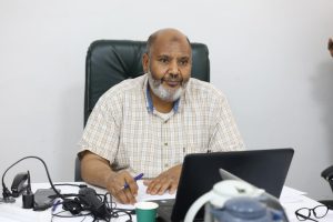 7F8C377A-837A-405D-A056-CD8789FDFF7C - جامعة سبها | Sebha University Middle-aged man with a gray beard sits at a desk in an office, writing with a blue pen beside a laptop.