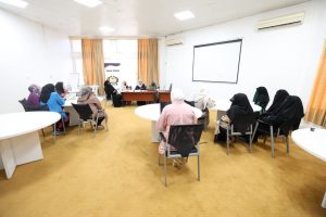 Group of women in hijabs seated around circular tables in a bright conference room, panel at the front with a projector screen behind them.