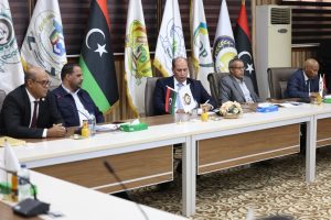 825E11F9-8918-4CC3-B020-AE905C08726D - جامعة سبها | Sebha University Five men in suits sit at a long conference table during a formal meeting with flags lined up behind them.