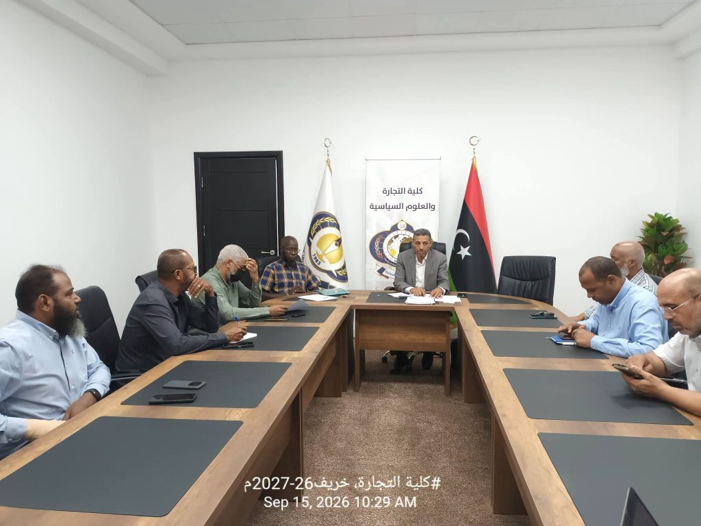 8435F9BA-38C3-47F7-B7B3-B80CE323B806 - جامعة سبها | Sebha University Business meeting in a bright conference room with several men seated around a long wooden table; flags and a banner with Arabic text behind the central figure.